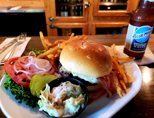 A delicious burger with bacon, lettuce, tomato, pickles, fries, coleslaw, and a bottle of Blue Moon beer on a wooden table.