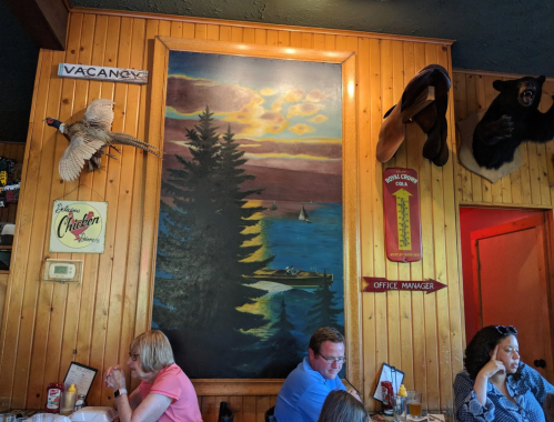 A cozy restaurant interior featuring a large sunset mural, wooden walls, and mounted animal decor. Diners are enjoying their meals.