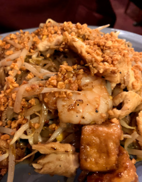 A plate of stir-fried noodles topped with chicken, shrimp, tofu, bean sprouts, and crushed peanuts.