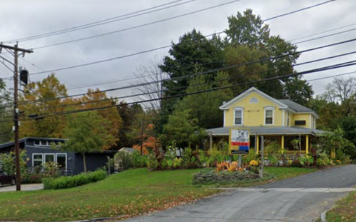 A yellow house with a porch and a sign, surrounded by greenery and trees, on a cloudy day. Power lines overhead.