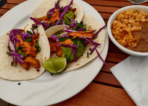 Two tacos with purple cabbage, guacamole, and sauce, served with rice and beans on a wooden table.