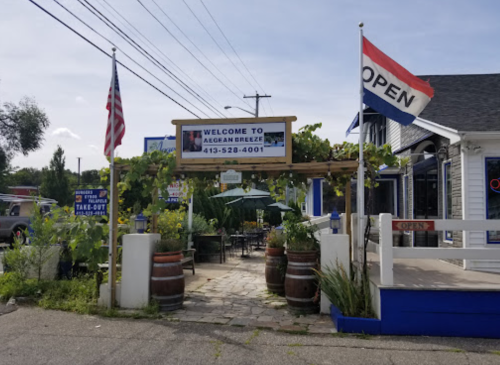 Entrance to Aegean Breeze restaurant with flags, a sign saying "Open," and outdoor seating surrounded by greenery.