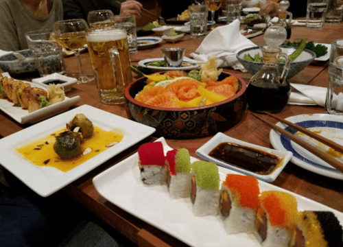 A table filled with various sushi rolls, sashimi, dipping sauces, and drinks, creating a vibrant dining scene.