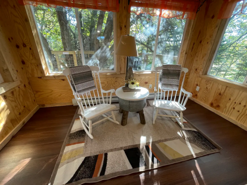 Cozy sunlit room with two rocking chairs, a small table, and large windows overlooking trees.