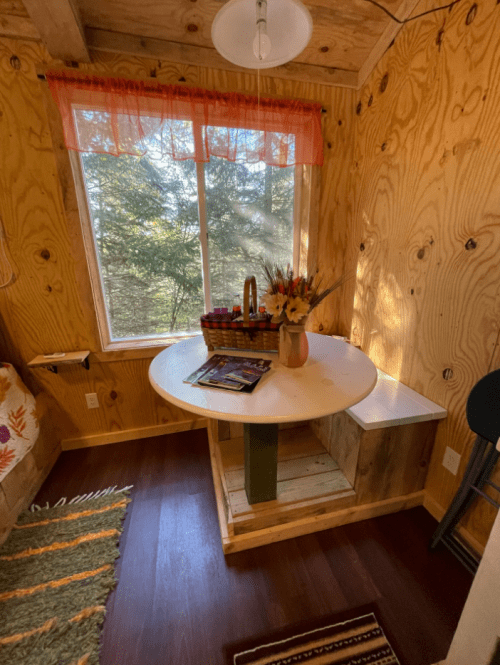 Cozy interior of a small cabin with a round table, a basket of flowers, and a window overlooking trees.