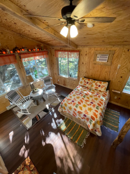 Cozy cabin interior with a bed, rocking chairs, and a small table, surrounded by wooden walls and natural light.