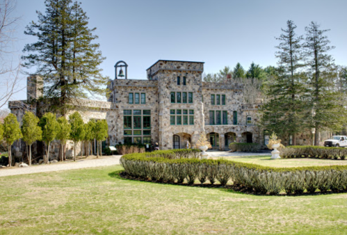 A large stone mansion surrounded by manicured lawns and trees, featuring a bell tower and elegant landscaping.