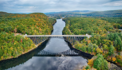 Aerial view of a bridge spanning a river, surrounded by lush green and autumn-colored trees under a cloudy sky.