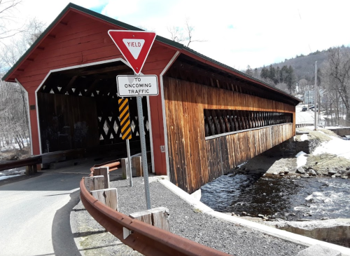 A red covered bridge with wooden siding, surrounded by snow and a road sign indicating to yield to oncoming traffic.