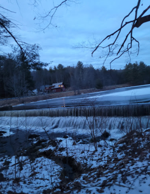 A serene winter scene featuring a partially frozen river, a cabin in the distance, and trees against a twilight sky.