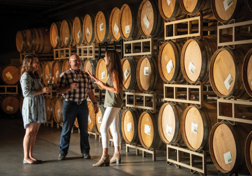 Two women and a man are chatting and holding wine glasses in a winery, surrounded by wooden barrels.