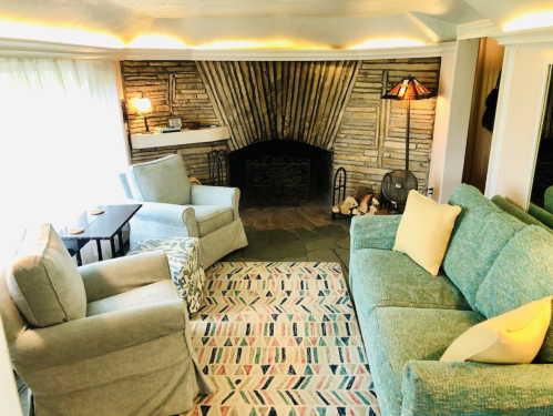 Cozy living room with two sofas, an armchair, a patterned rug, and a stone fireplace. Soft lighting creates a warm atmosphere.