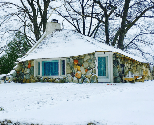 A unique stone cottage with a sloped roof, surrounded by snow and trees, creating a cozy winter scene.