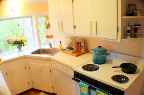 Bright kitchen with white cabinets, a sink, stove, and teal pot, featuring a window with flowers and colorful decor.