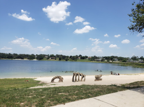 A serene lake scene with a sandy shore, grassy area, and unique stone sculptures under a blue sky with fluffy clouds.