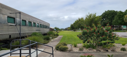 A vintage train car beside a grassy area with trees and shrubs under a cloudy sky.