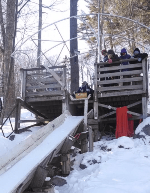 A snowy scene featuring a wooden play structure with a slide, where children are gathered on the upper level.