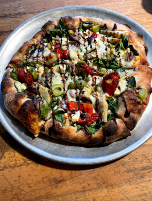 A freshly baked pizza topped with spinach, artichokes, roasted red peppers, olives, and drizzled with balsamic glaze.