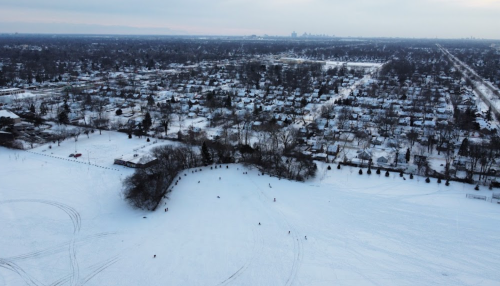 Aerial view of a snowy landscape with a park, trees, and houses in the background under a cloudy sky.