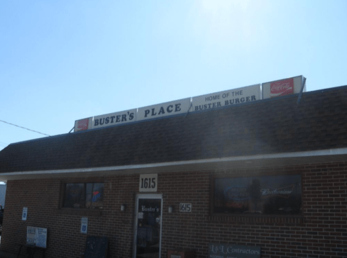 Exterior of Buster's Place, a restaurant featuring a sign that reads "Home of the Buster Burger." Clear blue sky above.