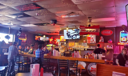 A lively bar interior with neon signs, a TV showing a motorcycle, and patrons enjoying food and drinks at the counter.