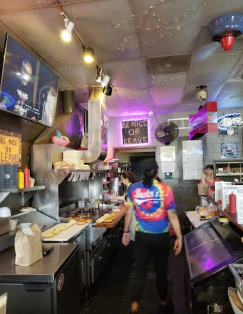 A busy diner kitchen with a chef in a colorful shirt, cooking and serving food, and a TV playing in the background.