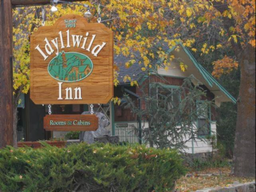Wooden sign for Idyllwild Inn, surrounded by autumn leaves and greenery, with a cozy cabin in the background.