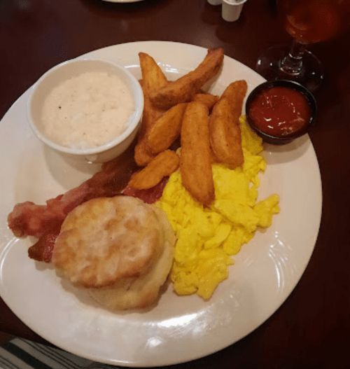 A plate of breakfast featuring scrambled eggs, bacon, a biscuit, potato wedges, and a side of rice with ketchup.