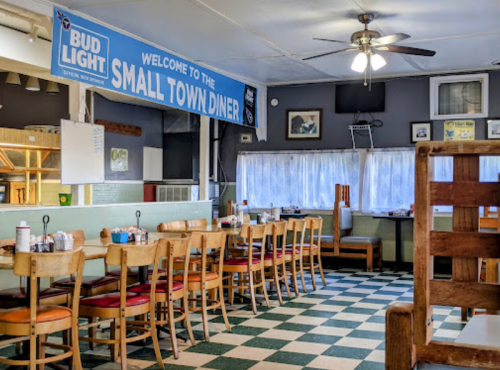 Interior of a diner with checkered floor, wooden tables, and a banner reading "Welcome to the Small Town Diner."