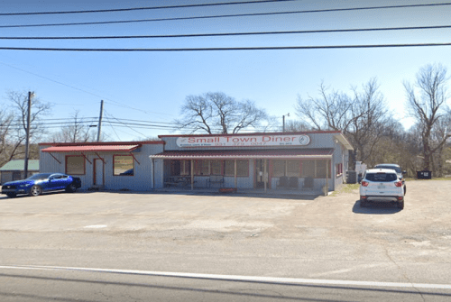 A small diner with a red roof and sign, surrounded by trees and parked cars on a sunny day.