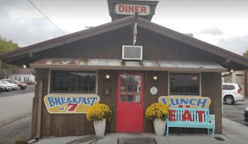 A cozy diner with a rustic exterior, featuring signs for breakfast and lunch, and colorful flowers by the entrance.