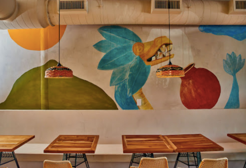 Colorful mural featuring a bird and abstract shapes on a restaurant wall, with wooden tables and hanging lights.