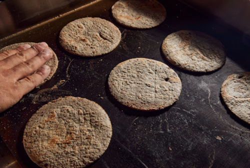 A hand touches a batch of round, rustic bread loaves on a baking tray, showcasing their textured surfaces.