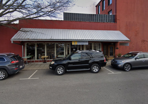 A storefront with a metal roof and large windows, flanked by parked cars on a city street.