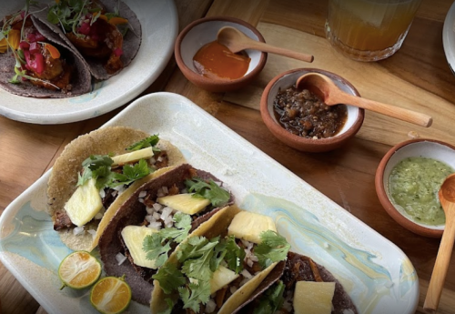 A plate of tacos with pineapple and cilantro, surrounded by small bowls of sauces and a drink on a wooden table.