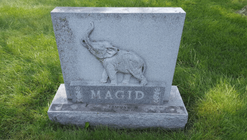 A gray granite gravestone featuring an elephant sculpture and the name "MAGID" engraved on it, surrounded by grass.