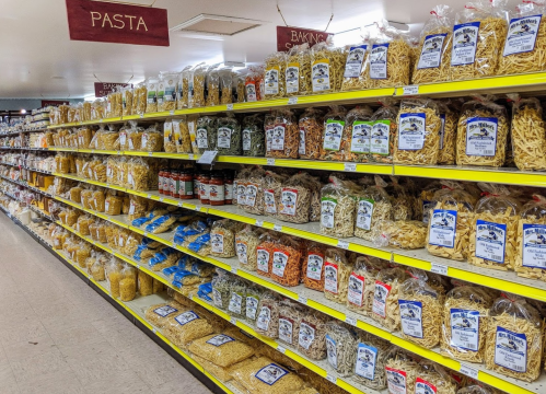 A grocery store aisle filled with various types of pasta and baking ingredients in clear bags on yellow shelves.