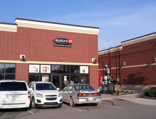 Exterior of a Burger 21 restaurant with a red brick facade and parked cars in front. Clear blue sky above.