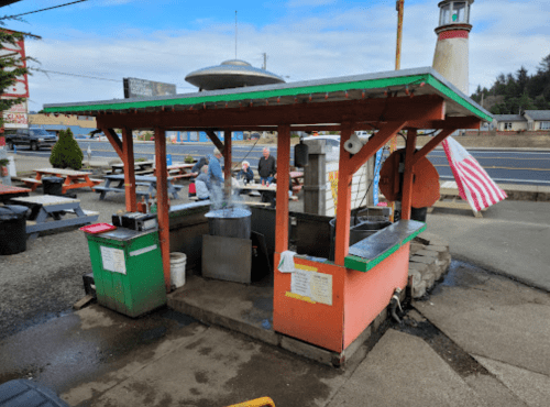 A colorful outdoor food stand with a steaming pot, picnic tables, and a lighthouse in the background.