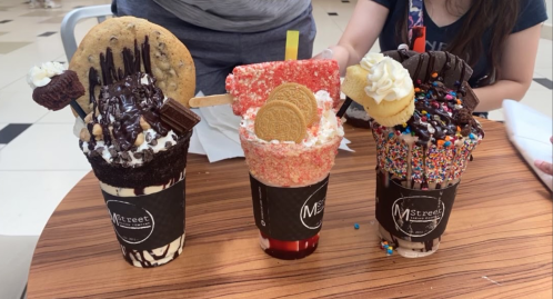 Three extravagant milkshakes topped with cookies, candy, and whipped cream, served in clear cups on a wooden table.