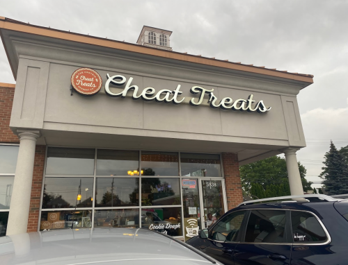 Exterior of a store named "Cheat Treats," featuring a sign and cloudy sky in the background. Cars parked in front.