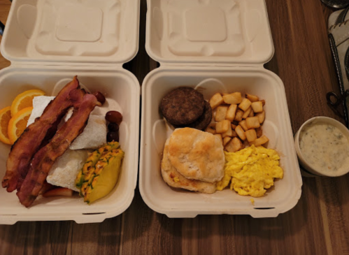 Two takeout containers with breakfast: bacon, eggs, biscuits, sausage, fruit, and potatoes.