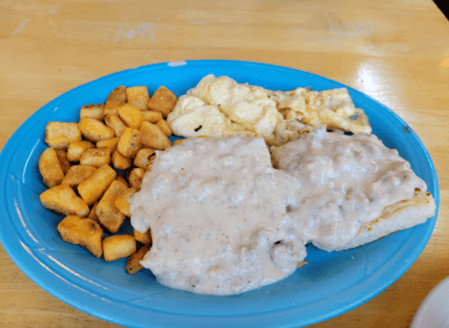 A blue plate with two biscuits topped with gravy, scrambled eggs, and seasoned potato cubes.