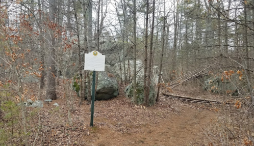 A wooded area with a historical marker sign and large rocks, surrounded by trees and fallen leaves.