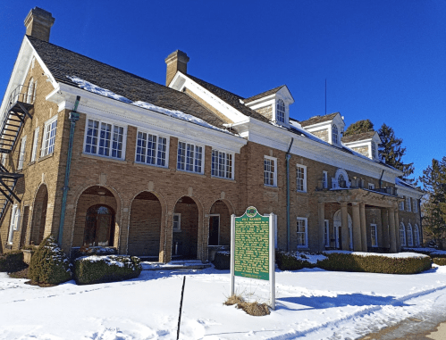 Historic brick building with arched windows, surrounded by snow and greenery, under a clear blue sky.