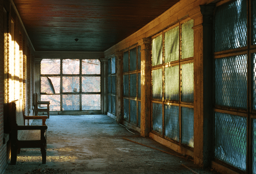 A dimly lit corridor with large windows, wooden benches, and a weathered floor, creating a nostalgic atmosphere.