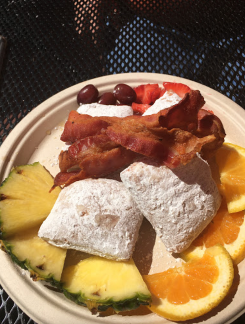A plate with powdered sugar pastries, bacon, and fresh fruit including pineapple, orange, strawberries, and grapes.