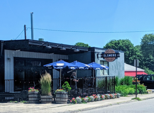 Exterior of Elsmere restaurant with outdoor seating, umbrellas, and colorful flower planters on a sunny day.