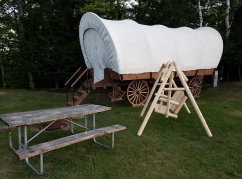 A covered wagon on wheels beside a wooden swing and a picnic table in a grassy area surrounded by trees.