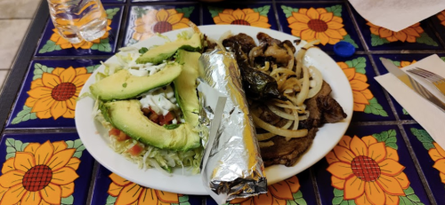 A plate with grilled meat, onions, avocado, salad, and a foil-wrapped burrito on a colorful floral table.